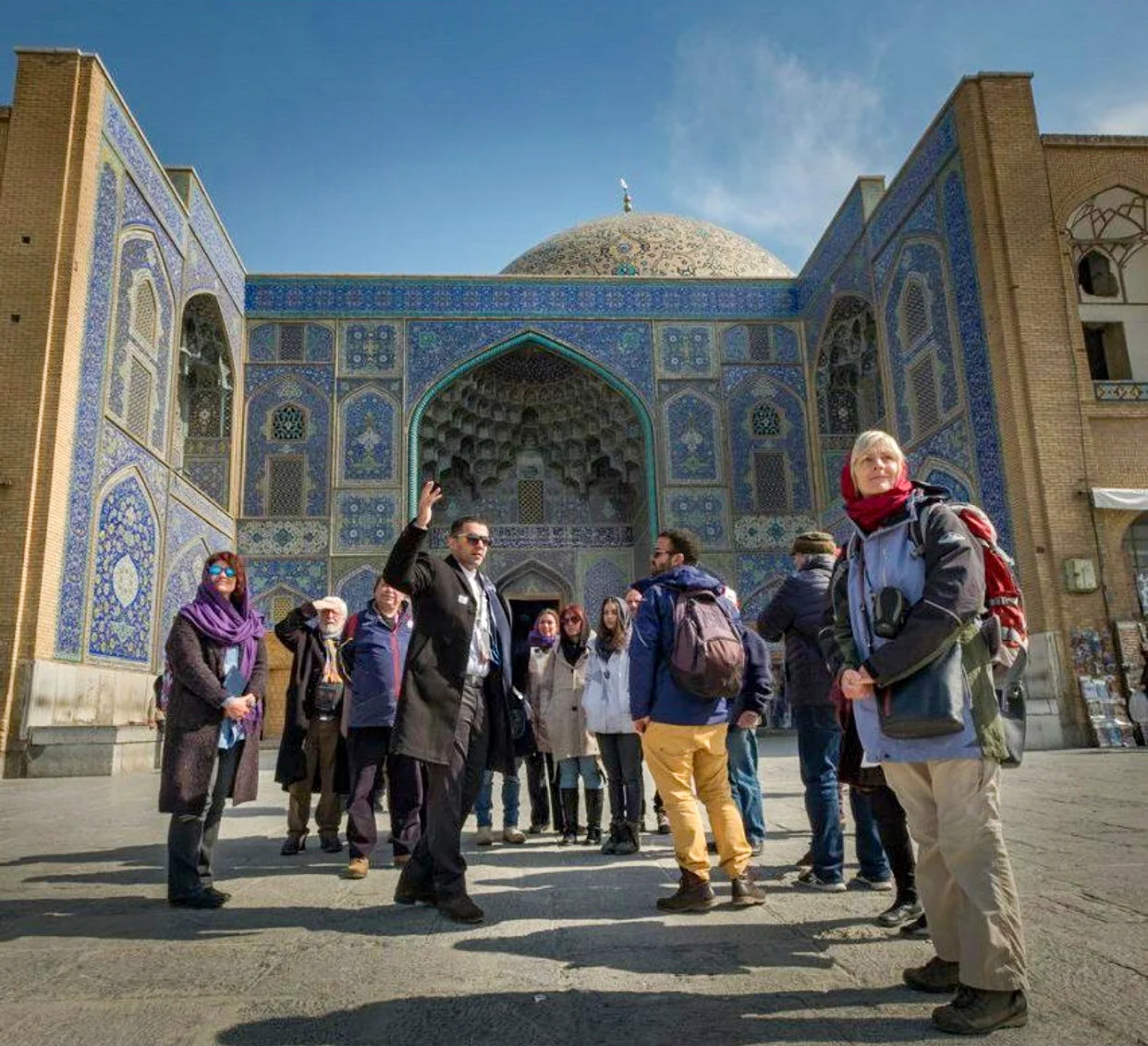 A local guide in Iran gestures toward the historic Sheikh lotfollah Mosque in Naqsh-e Jahan Square, Isfahan, while leading a group of international tourists on a cloudy day.