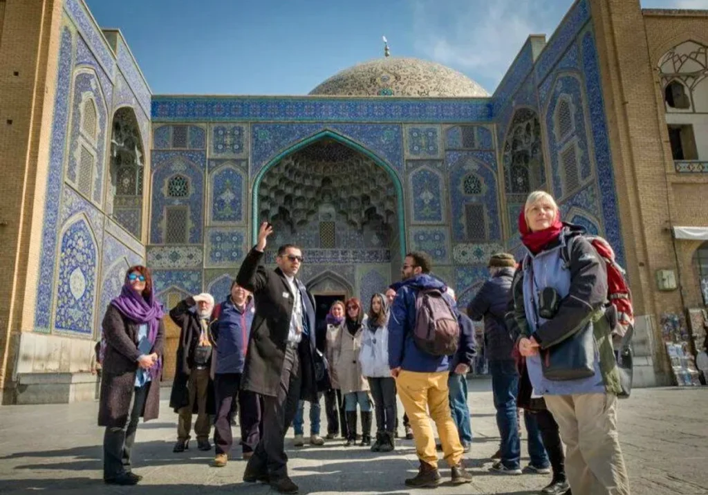 A local guide in Iran gestures toward the historic Sheikh lotfollah Mosque in Naqsh-e Jahan Square, Isfahan, while leading a group of international tourists on a cloudy day.